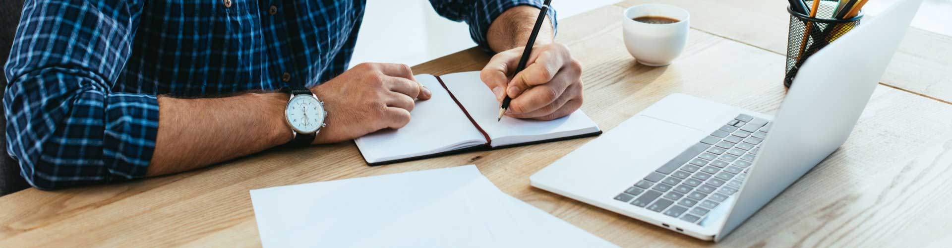 Man writing in a notebook in front of a laptop